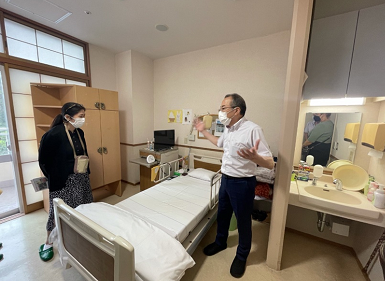 A man and woman speak across a medical bed in a hospital room.