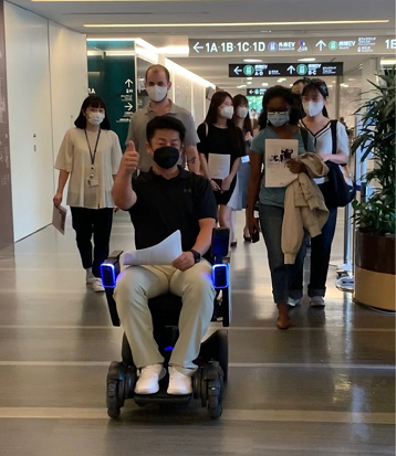 A man rides an automatic wheelchair with students following behind